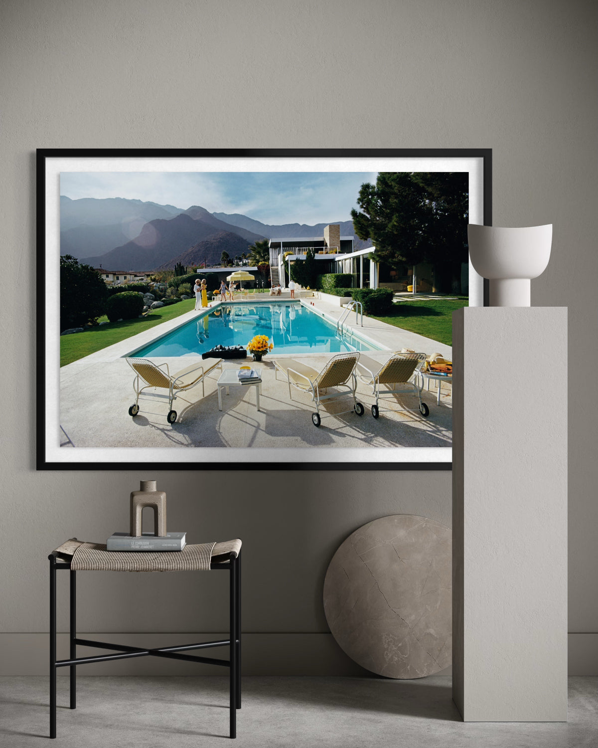 A framed art print depicting a luxurious outdoor poolside scene with mountains in the background, displayed on a gray wall in a modern styled room setting.