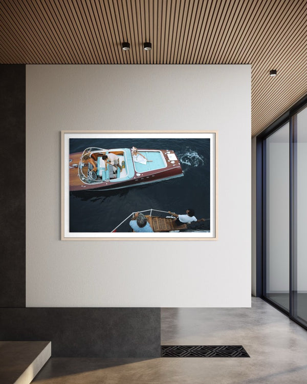 A framed art print depicting an aerial view of a red and blue motorboat on dark water, displayed on a white wall in a modern interior setting with a slatted wood ceiling. The Picturalist Wall Art and Photography for Designed Interiors. Slim Aarons Collection, Riva Boat. Getty Images Gallery. 