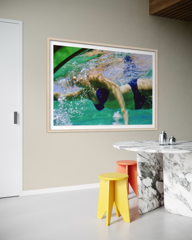 A framed square art print depicting an underwater swim scene with turquoise water and a swimmer in dark blue trunks, hung on a beige wall above a marble table with colorful stools in a modern room. The Picturalist Wall Art and Photography for Designed Interiors. Slim Aarons Collection, Poolside. Getty Images Gallery. 