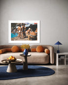 A framed art print depicting a vintage beach poolside scene with people in swimwear, displayed on a light gray wall above a beige sofa in a modern living room setting. The Picturalist Wall Art and Photography for Designed Interiors. Slim Aarons Collection, Poolside. Getty Images Gallery. 