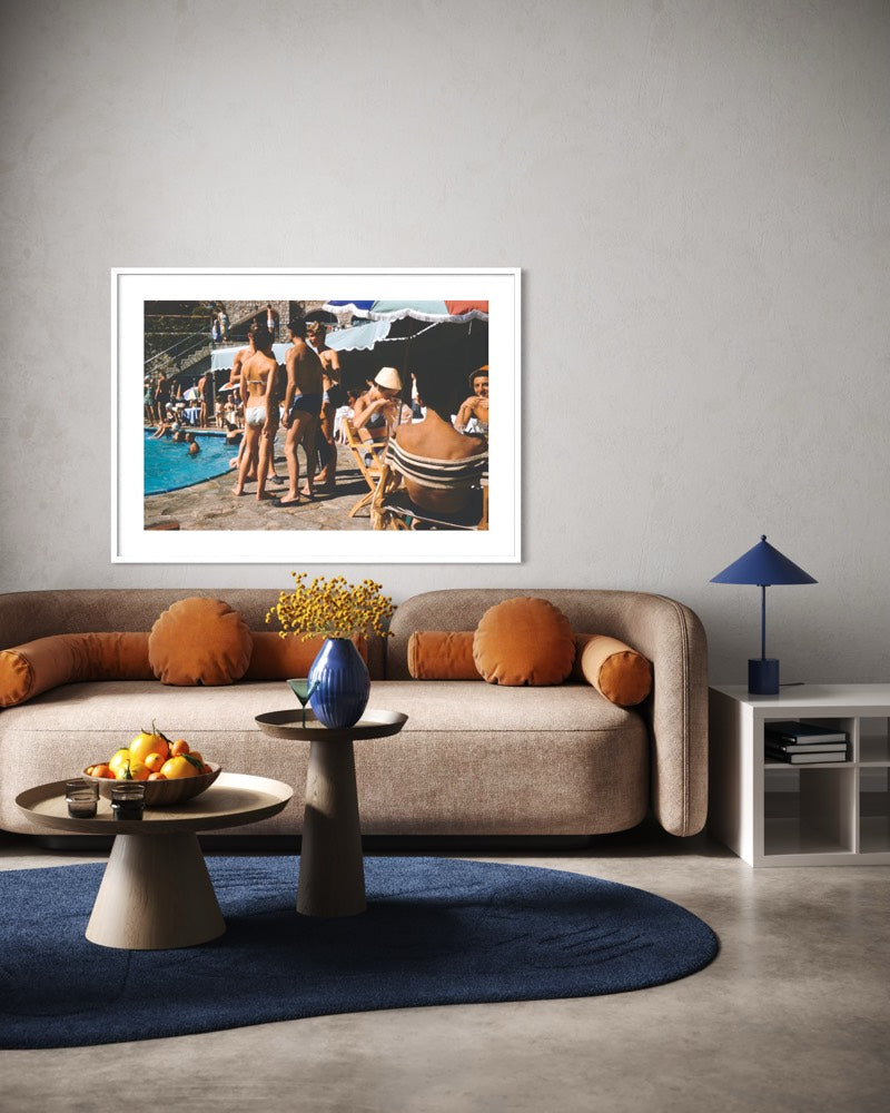 A framed art print depicting a vintage beach poolside scene with people in swimwear, displayed on a light gray wall above a beige sofa in a modern living room setting. The Picturalist Wall Art and Photography for Designed Interiors. Slim Aarons Collection, Poolside. Getty Images Gallery. 