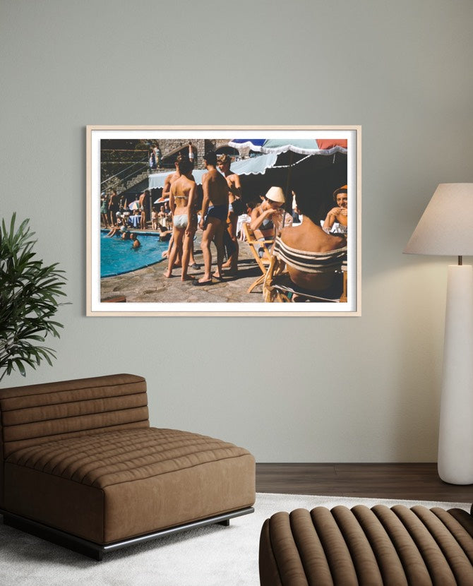 A framed art print depicting a vintage poolside scene with people, displayed on a light gray wall in a modern living room setting with a brown leather lounge chair and a floor lamp. The Picturalist Wall Art and Photography for Designed Interiors. Slim Aarons Collection, Poolside. Getty Images Gallery. 