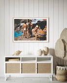 A framed art print depicting a vintage beach pool party scene, styled on a white paneled wall above a white sideboard cabinet with woven cane doors, flanked by decorative ceramic vases and a potted plant. The Picturalist Wall Art and Photography for Designed Interiors. Slim Aarons Collection, Poolside. Getty Images Gallery. 