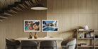 Two framed art prints depicting poolside scenes, displayed side-by-side on a light wood paneled wall in a modern dining room setting. The Picturalist Wall Art and Photography for Designed Interiors. Slim Aarons Collection, Poolside. Getty Images Gallery. 
