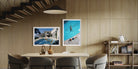 Two framed art prints hanging on a light wood-paneled wall in a modern dining room setting. The left print shows a modern villa with a pool, and the right print shows an aerial view of a swimmer in a bright blue pool. The Picturalist Wall Art and Photography for Designed Interiors. Slim Aarons Collection, Poolside. Getty Images Gallery. 