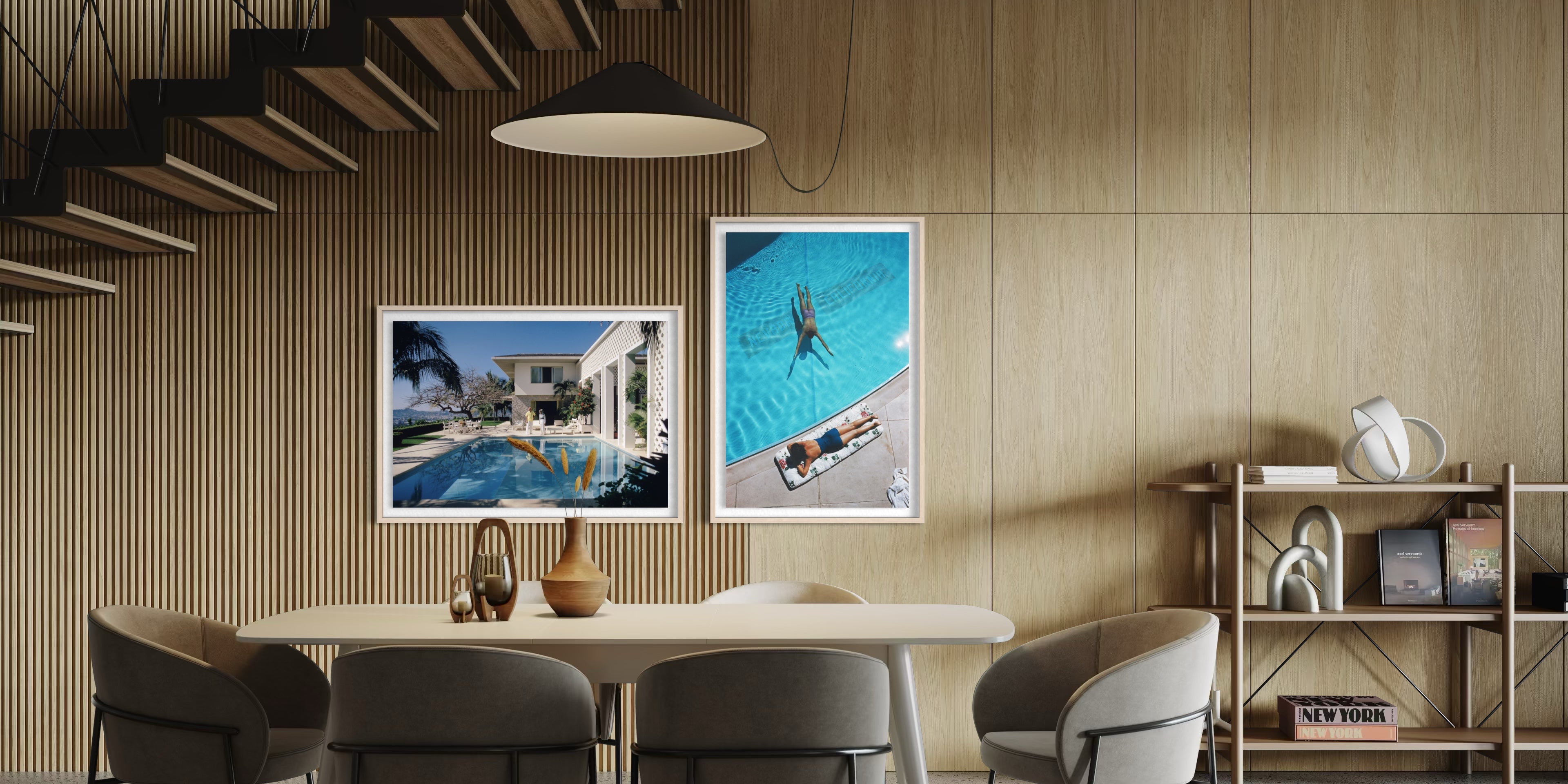 Two framed art prints hanging on a light wood-paneled wall in a modern dining room setting. The left print shows a modern villa with a pool, and the right print shows an aerial view of a swimmer in a bright blue pool. The Picturalist Wall Art and Photography for Designed Interiors. Slim Aarons Collection, Poolside. Getty Images Gallery. 