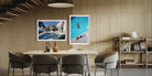Two framed art prints hanging on a light wood slatted wall above a dining table. The left print shows a poolside villa scene, and the right print shows an aerial view of a swimmer in a bright blue pool. The Picturalist Wall Art and Photography for Designed Interiors. Slim Aarons Collection, Poolside. Getty Images Gallery. 