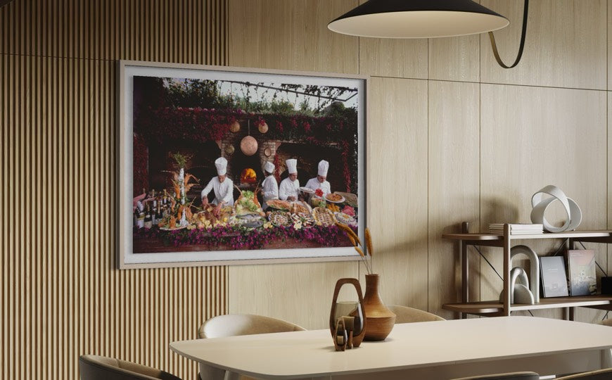 A framed art print depicting chefs preparing food hangs on a light wood slatted wall in a modern dining room setting with a table, chairs, and a pendant light. The Picturalist Wall Art and Photography for Designed Interiors. Slim Aarons Collection, Poolside. Getty Images Gallery. 