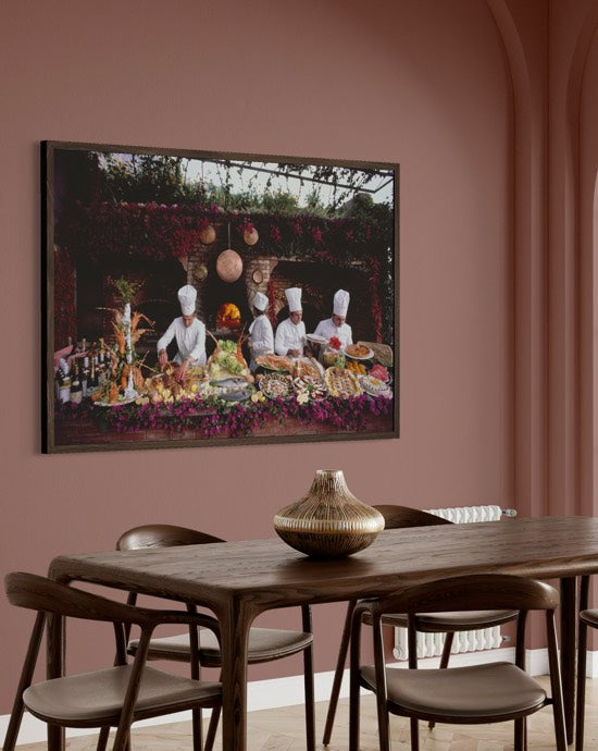 A framed art print depicting chefs serving food hangs on a dusty rose wall above a wooden dining table with chairs. The Picturalist Wall Art and Photography for Designed Interiors. Slim Aarons Collection, Poolside. Getty Images Gallery. 