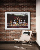 A framed art print depicting a restaurant scene with chefs and food, displayed on a brick wall above a modern beige lounge chair. The Picturalist Wall Art and Photography for Designed Interiors. Slim Aarons Collection, Poolside. Getty Images Gallery. 