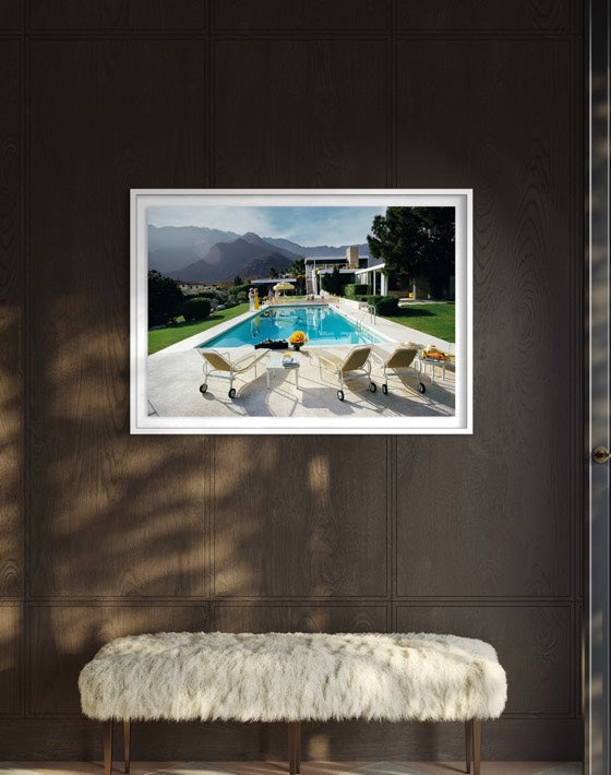 A framed art print depicting a luxurious outdoor poolside scene with mountains in the background, displayed on a dark wood-paneled wall above a white faux-fur bench. The Picturalist Wall Art and Photography for Designed Interiors. Slim Aarons Collection, Poolside. Getty Images Gallery. 