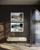 Two framed landscape art prints are displayed on a dark brown paneled wall above a white faux-fur bench. The prints depict outdoor scenes with mountains, a pool, and a beach. The Picturalist Wall Art and Photography for Designed Interiors. Slim Aarons Collection, Poolside. Getty Images Gallery. 