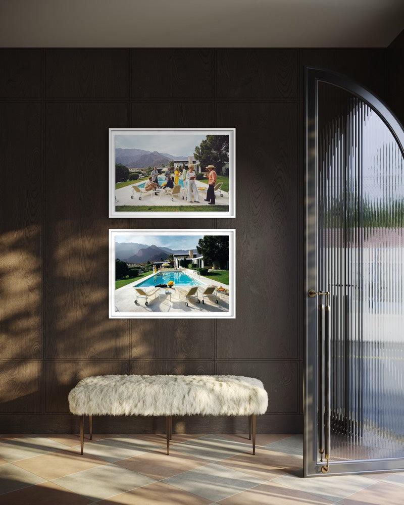 Two framed landscape art prints are displayed on a dark brown paneled wall above a white faux-fur bench. The prints depict outdoor scenes with mountains, a pool, and a beach. The Picturalist Wall Art and Photography for Designed Interiors. Slim Aarons Collection, Poolside. Getty Images Gallery. 