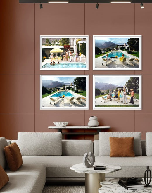 A modern living room setting featuring a wall-mounted four-panel framed art print set. The prints showcase outdoor poolside scenes with people, set against a warm brown paneled wall above a contemporary light gray sectional sofa. The Picturalist Wall Art and Photography for Designed Interiors. Slim Aarons Collection, Poolside. Getty Images Gallery. 