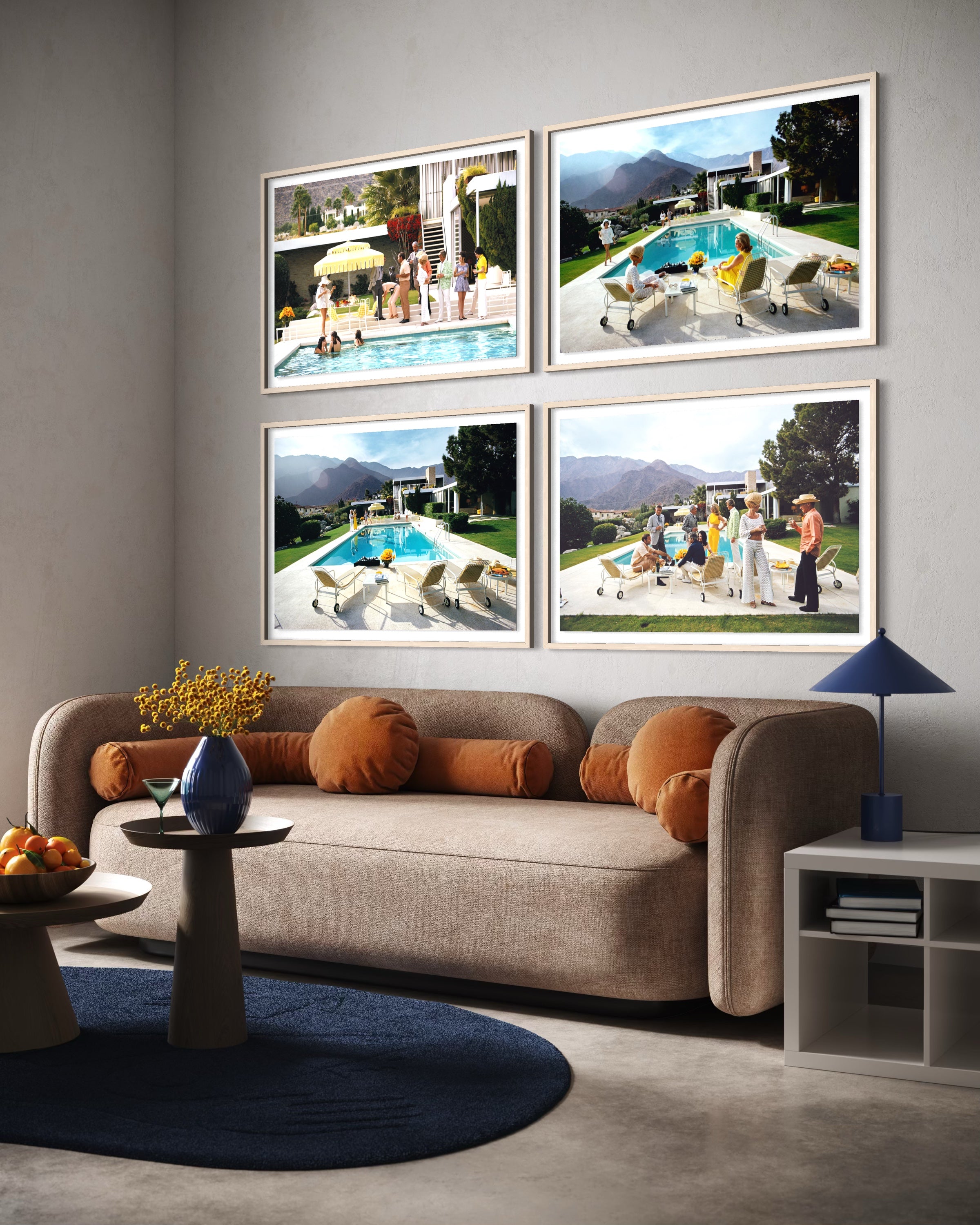 A styled living room interior featuring a beige sofa with orange cushions, a round blue rug, and a side table. Above the sofa, four framed photographic prints depicting poolside scenes are mounted on a light gray wall. The Picturalist Wall Art and Photography for Designed Interiors. Slim Aarons Collection, Poolside. Getty Images Gallery. 