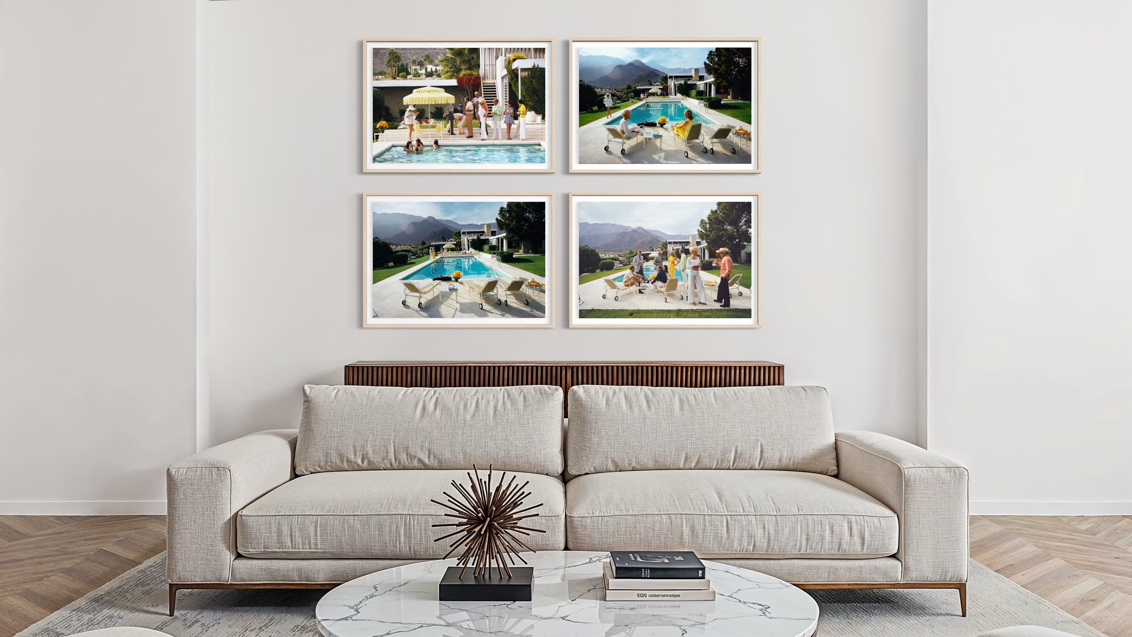 A modern living room setting featuring a light beige sofa and a round marble coffee table, with a wall-mounted four-panel framed print collage above the couch. The print set displays four different outdoor poolside and mountain landscape photographs in white frames. The Picturalist Wall Art and Photography for Designed Interiors. Slim Aarons Collection, Poolside. Getty Images Gallery. 