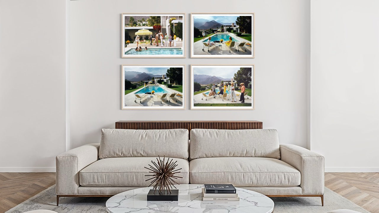 A modern living room setting featuring a light beige sofa and a round marble coffee table, with a wall-mounted four-panel framed print set above the couch. The print set displays four different outdoor poolside and mountain landscape photographs in white frames. The Picturalist Wall Art and Photography for Designed Interiors. Slim Aarons Collection, Poolside. Getty Images Gallery. 
