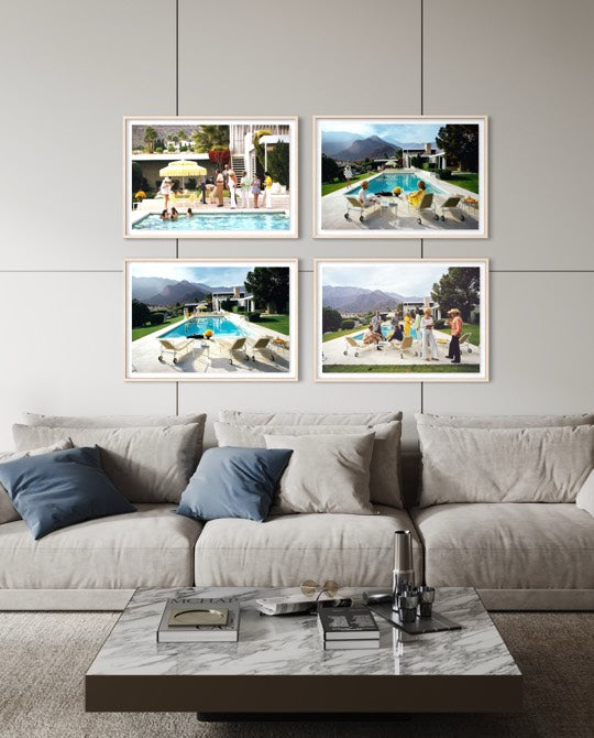 A styled living room scene showing a light beige sofa with blue and beige pillows, a marble coffee table, and four framed photographic prints hanging on the wall above the couch. The prints depict poolside scenes with people and mountains. The Picturalist Wall Art and Photography for Designed Interiors. Slim Aarons Collection, Poolside. Getty Images Gallery. 
