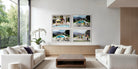 A modern living room with two white sofas and a wooden coffee table, featuring a wall-mounted four-panel framed print set above a low wooden console. The artwork consists of four square color photographs showing poolside scenes with people and outdoor landscapes. The Picturalist Wall Art and Photography for Designed Interiors. Slim Aarons Collection, Poolside. Getty Images Gallery. 