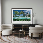 A framed art print depicting a tropical beach resort with palm trees, a swimming pool, and a thatched-roof structure, displayed on a light gray wall above a black console table in a modern living room setting. The Picturalist Wall Art and Photography for Designed Interiors. Slim Aarons Collection, Poolside. Getty Images Gallery. 