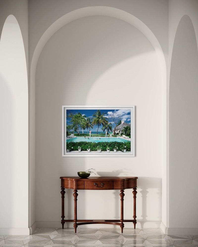 A framed tropical beach and poolside landscape print hangs on a white wall in an arched interior setting above a wooden console table. The Picturalist Wall Art and Photography for Designed Interiors. Slim Aarons Collection, Poolside. Getty Images Gallery. 