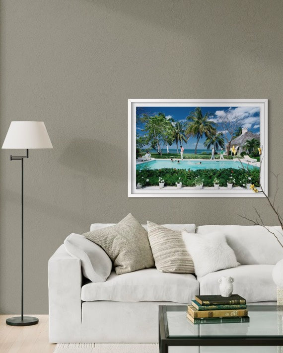 A framed tropical beach scene wall art print hangs above a white sofa in a modern living room setting with a floor lamp and glass coffee table. The Picturalist Wall Art and Photography for Designed Interiors. Slim Aarons Collection, Poolside. Getty Images Gallery. 