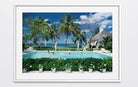 A framed art print depicting a tropical resort scene with a swimming pool, palm trees, ocean, and a thatched-roof building, shown against a light gray background. The Picturalist Wall Art and Photography for Designed Interiors. Slim Aarons Collection, Poolside. Getty Images Gallery. 