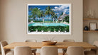 A framed tropical beach resort painting hanging on a beige wall above a wooden dining table with chairs. The artwork depicts palm trees, a swimming pool, beach umbrellas, and a blue sky. The Picturalist Wall Art and Photography for Designed Interiors. Slim Aarons Collection, Poolside. Getty Images Gallery. 