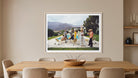 A framed art print depicting a group of people relaxing by a pool in a sunny outdoor setting, displayed on a beige wall above a wooden dining table in a modern dining room. The Picturalist Wall Art and Photography for Designed Interiors. Slim Aarons Collection, Poolside. Getty Images Gallery. 