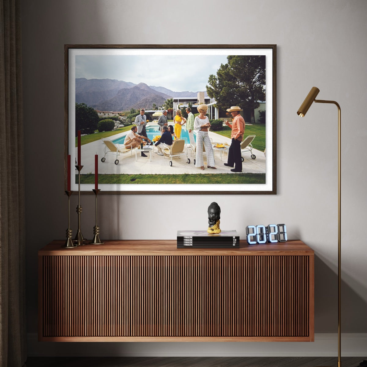 A framed art print depicting a classic mid-century modern pool party scene, displayed on a neutral gray wall above a wooden console table in a styled interior setting. The Picturalist Wall Art and Photography for Designed Interiors. Slim Aarons Collection, Poolside. Getty Images Gallery. 