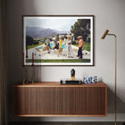 A framed art print depicting a classic mid-century modern pool party scene, displayed on a neutral gray wall above a wooden console table in a styled interior setting. The Picturalist Wall Art and Photography for Designed Interiors. Slim Aarons Collection, Poolside. Getty Images Gallery. 