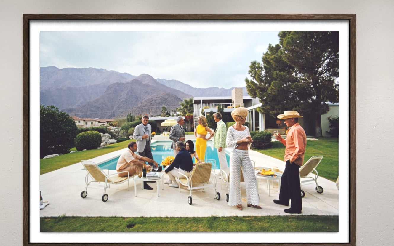 A framed photographic print of a 1960s-style poolside social gathering at a modern house with mountains in the background, displayed on a light gray wall. The Picturalist Wall Art and Photography for Designed Interiors. Slim Aarons Collection, Poolside. Getty Images Gallery. 