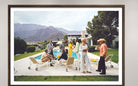 A framed photographic print of a 1960s-style poolside social gathering at a modern house with mountains in the background, displayed on a light gray wall. The Picturalist Wall Art and Photography for Designed Interiors. Slim Aarons Collection, Poolside. Getty Images Gallery. 