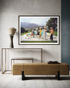 A framed art print depicting a vintage outdoor scene with people and a pool, styled in a modern interior setting above a textured bench and console table. The Picturalist Wall Art and Photography for Designed Interiors. Slim Aarons Collection, Poolside. Getty Images Gallery. 