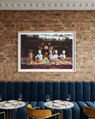 A framed art print depicting three chefs at a buffet table, displayed on a brick wall above a blue velvet restaurant booth. The Picturalist Wall Art and Photography for Designed Interiors. Getty Images Gallery. 