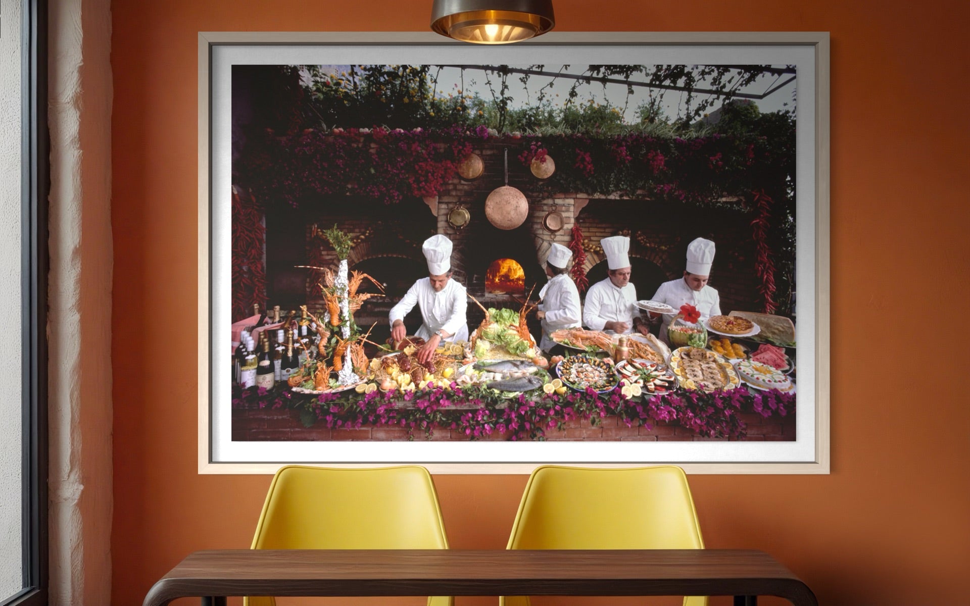 A large framed photographic print hangs on an orange-brown wall above a table with two yellow chairs. The print shows chefs preparing food at an outdoor dining table surrounded by abundant dishes and hanging lanterns. The Picturalist Wall Art and Photography for Designed Interiors. Getty Images Gallery. 