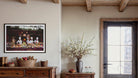 A framed art print depicting a rustic outdoor picnic scene with figures and food, displayed on a beige wall in a styled room with a wooden dresser, a vase of white blossoms, and a dark wood door. The Picturalist Wall Art and Photography for Designed Interiors. Getty Images Gallery. 