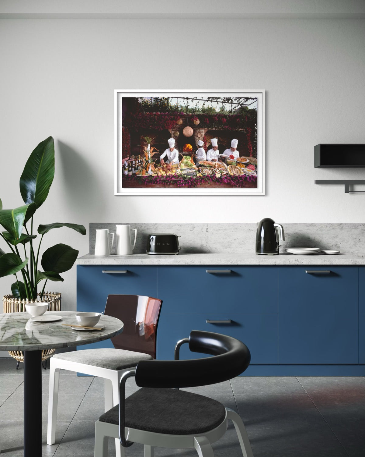 A framed art print depicting a dinner scene with chefs, displayed on a light gray wall above a modern kitchen with blue cabinets and a marble countertop. The Picturalist Wall Art and Photography for Designed Interiors. Getty Images Gallery. 