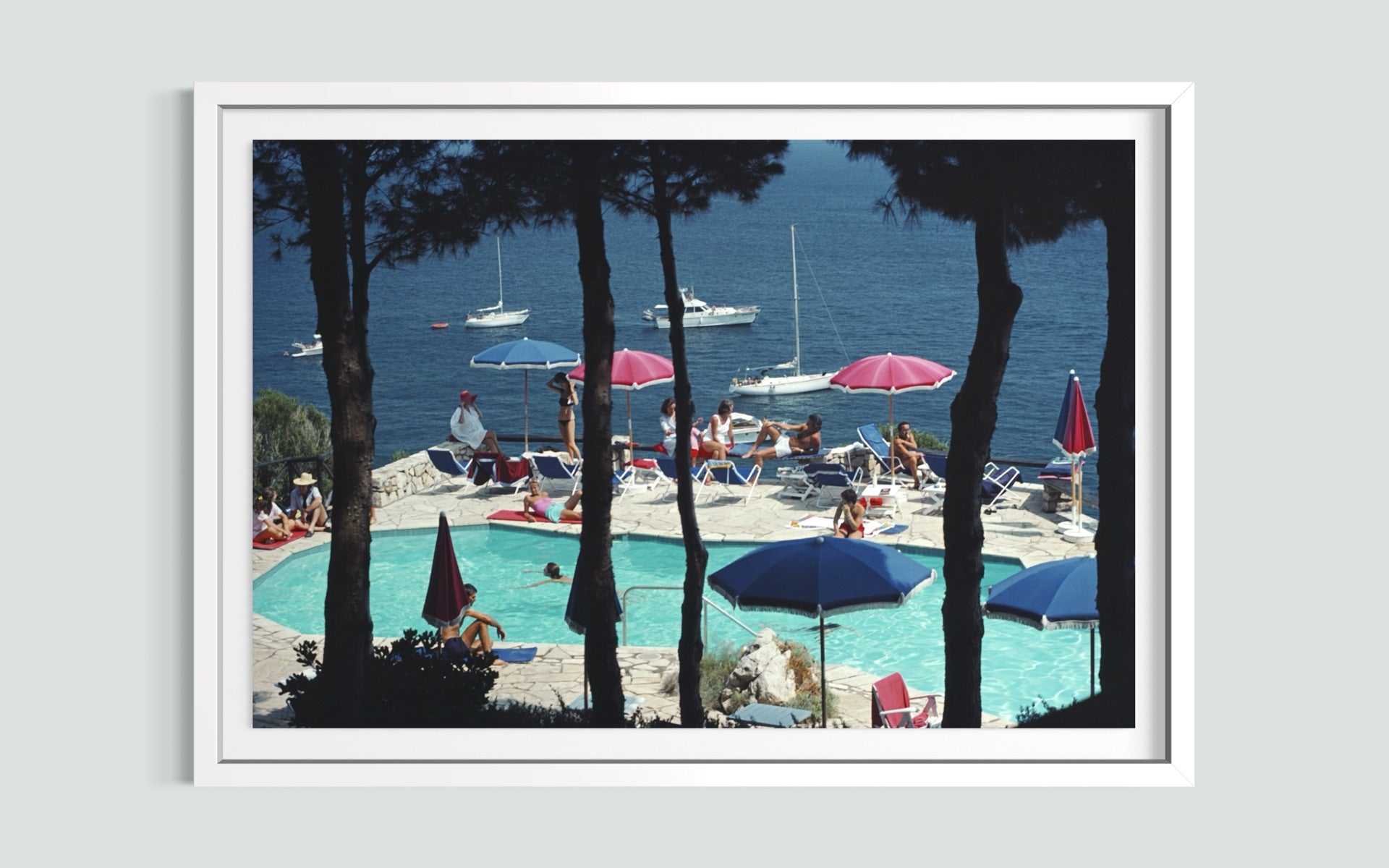 A framed art print depicting a scenic poolside view with sailboats on the blue sea, framed by dark pine tree silhouettes, and presented on a light gray wall.