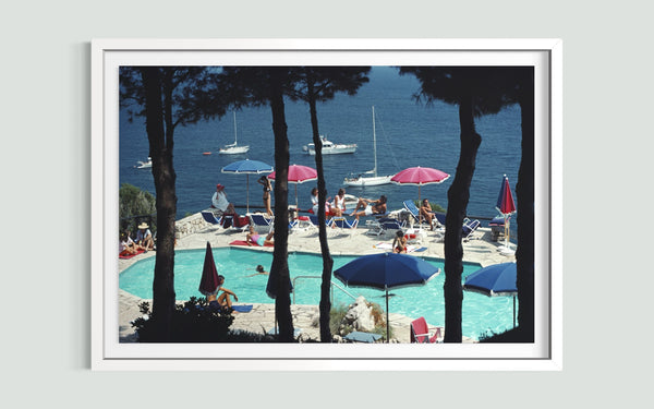 A framed art print depicting a scenic poolside view with sailboats on the blue sea, framed by dark pine tree silhouettes, and presented on a light gray wall.