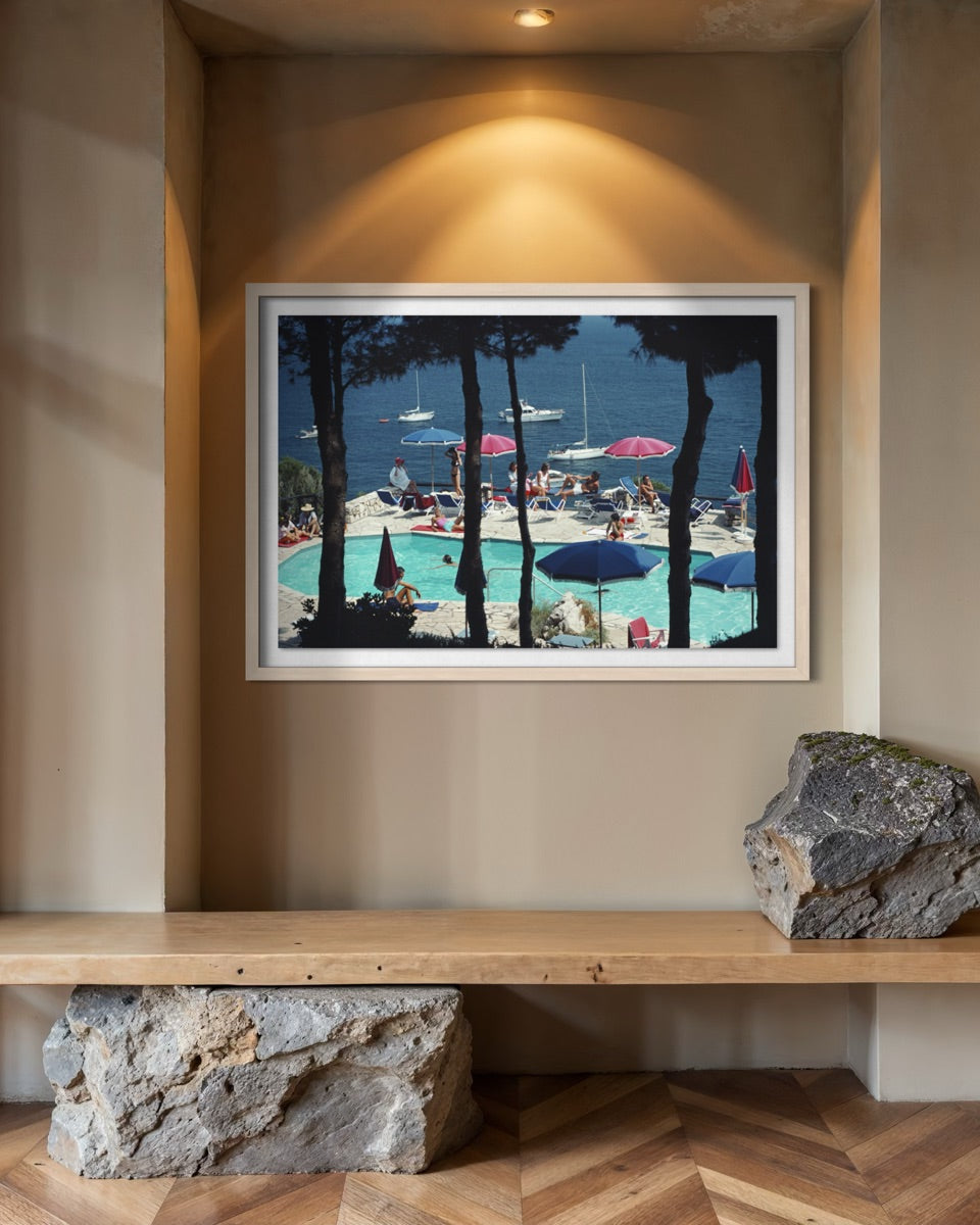A framed art print depicting a sunny poolside scene with blue water, umbrellas, and tall dark trees, displayed on a beige wall above a wooden bench with decorative rocks.