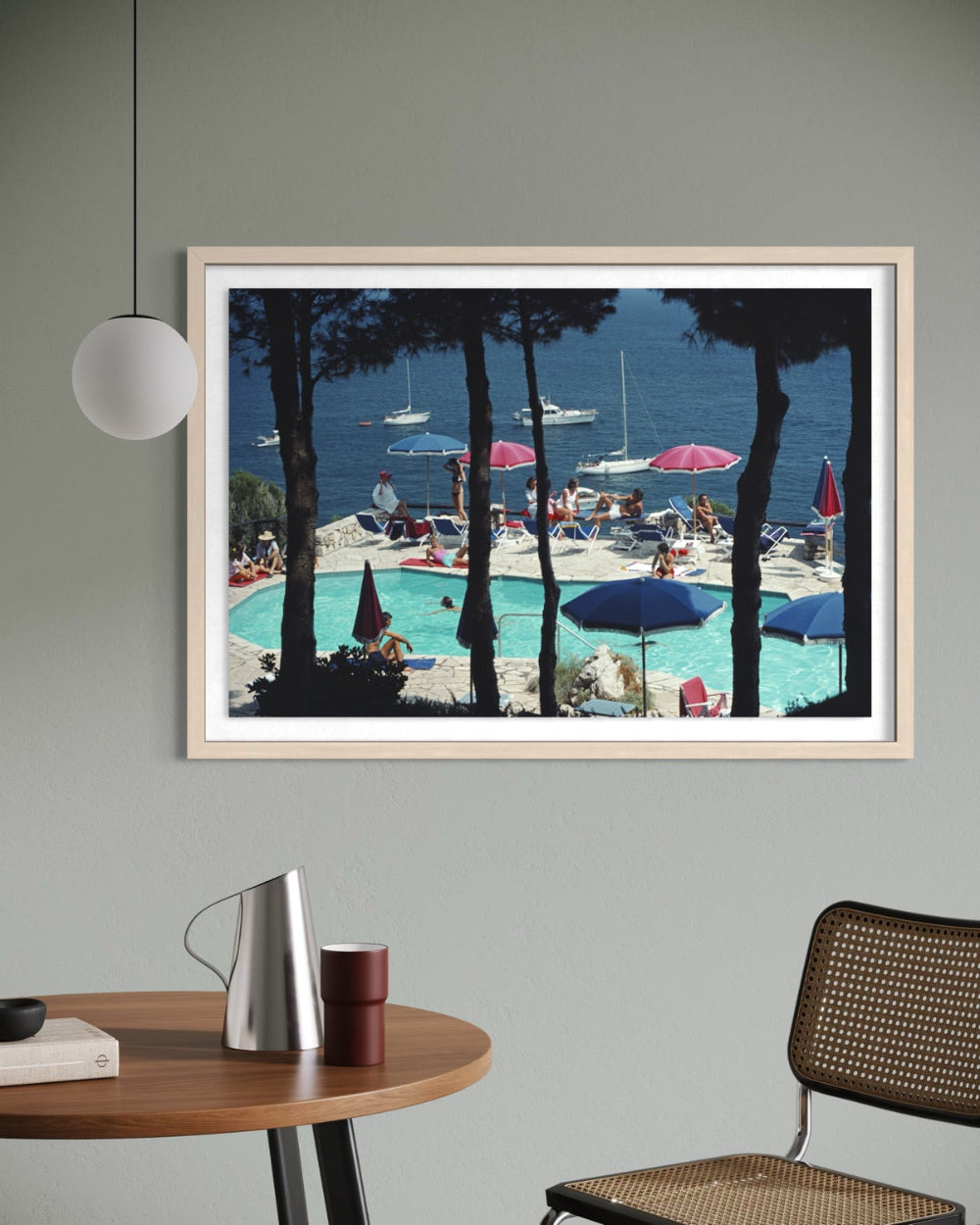 A framed art print depicting a sunny seaside poolside scene with blue water, white sailboats, and lounge chairs framed by dark pine trees, hung on a gray wall above a wooden table in a modern room setting.