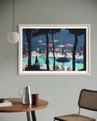 A framed art print depicting a sunny seaside poolside scene with blue water, white sailboats, and lounge chairs framed by dark pine trees, hung on a gray wall above a wooden table in a modern room setting.