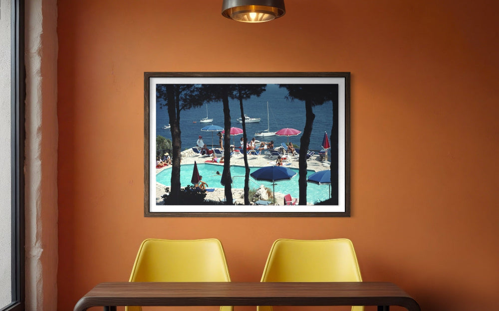 A framed art print depicting a sunny poolside beach scene with tall pine trees, blue umbrellas, and boats on the water, hung on an orange wall above a table with two yellow chairs.