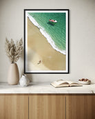 A framed vertical art print of an aerial beach scene with turquoise water, white waves, and a sandy shore, displayed on a light beige wall above a marble countertop with decorative vases, an open book, and a small bowl.