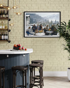 A framed horizontal landscape photograph print hanging on a light beige brick wall above a modern kitchen island with dark wood stools. The print depicts a winter mountain village scene with a group of people dining outdoors.