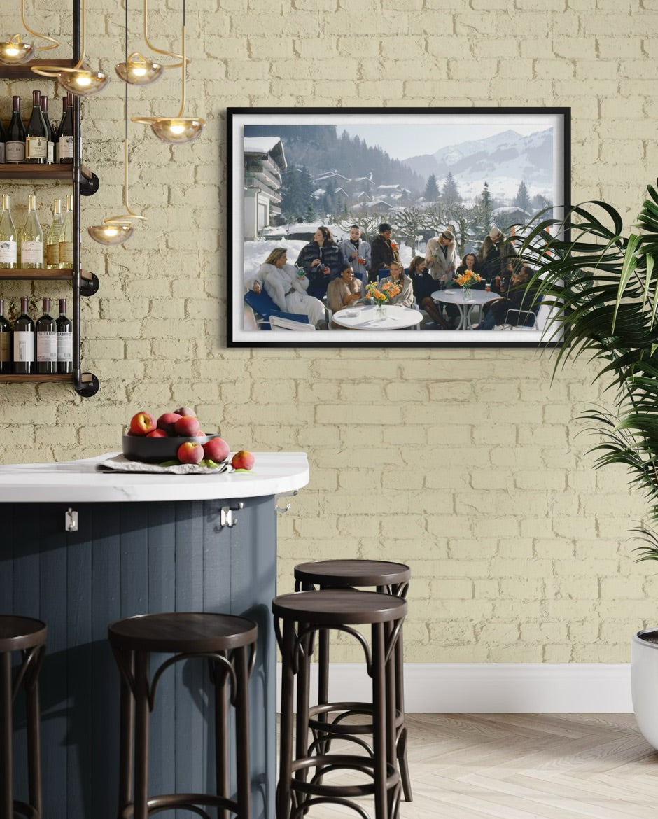 A framed horizontal landscape photograph print hanging on a light beige brick wall above a modern kitchen island with dark wood stools. The print depicts a winter mountain village scene with a group of people dining outdoors.