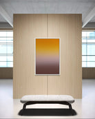 A vertical framed art print featuring a smooth yellow-to-brown gradient, displayed on a light wood panel wall above a modern white bench in a minimalist interior setting.