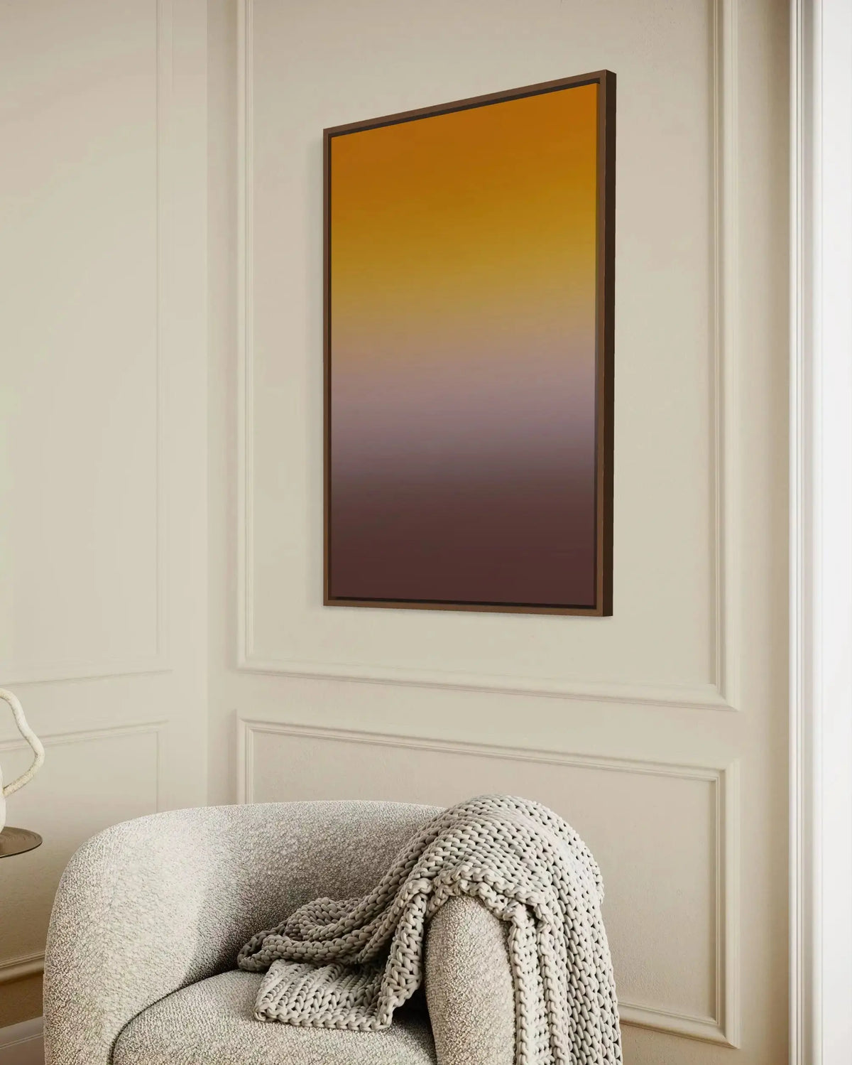 A framed vertical wall art print with a smooth gradient from golden yellow at the top to deep brown at the bottom, hanging on a cream paneled wall above a textured armchair with a knit throw blanket.