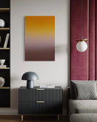 A vertical canvas wall art print featuring a smooth gradient from golden yellow at the top to warm brown at the bottom, displayed in a modern living room setting above a black ribbed sideboard.